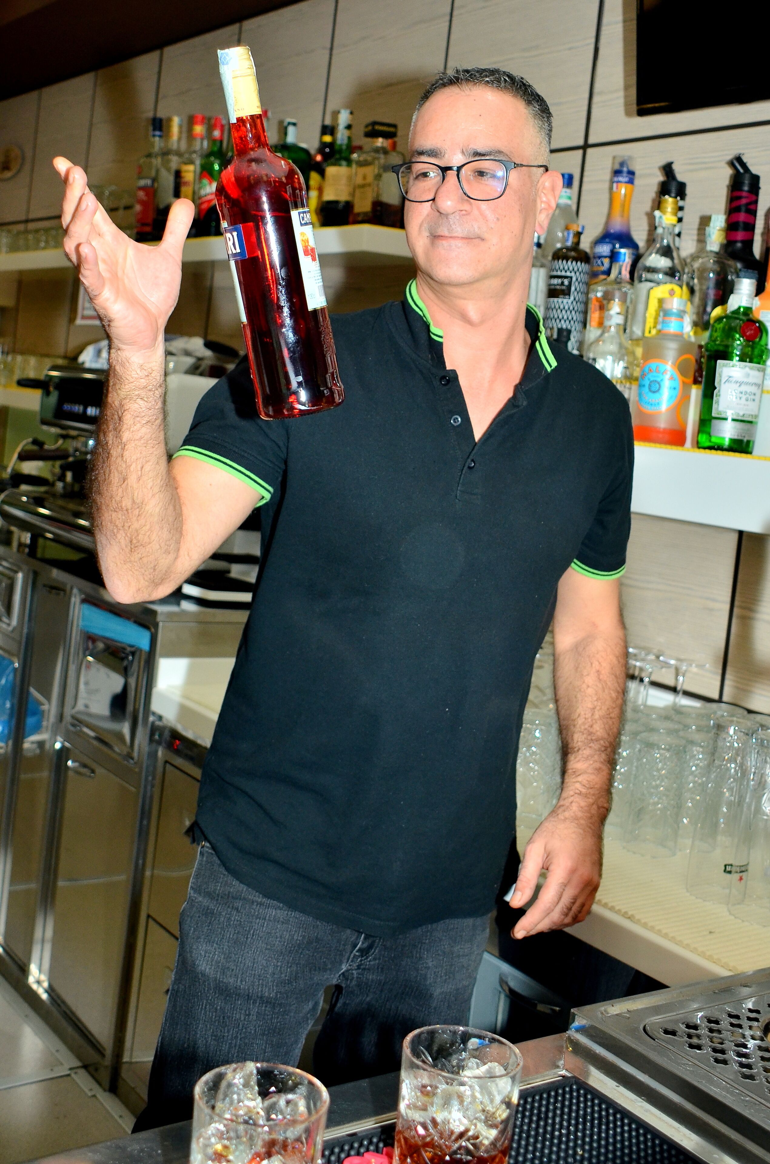 Una zona de bar con un hombre sosteniendo una botella, vasos sobre la barra y varias botellas al fondo.