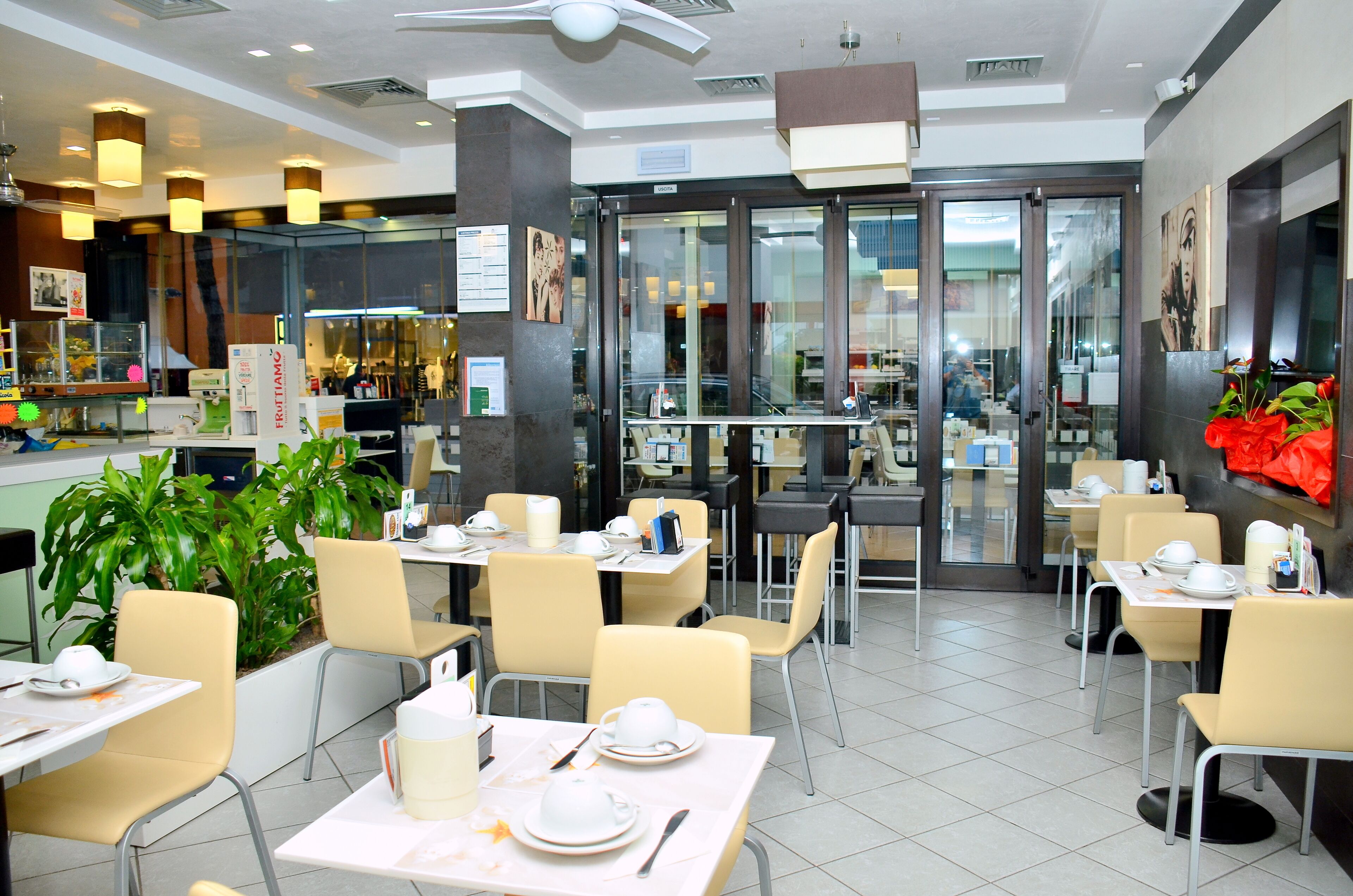 A modern restaurant with multiple dining tables and chairs, a glass entrance, and indoor plants.