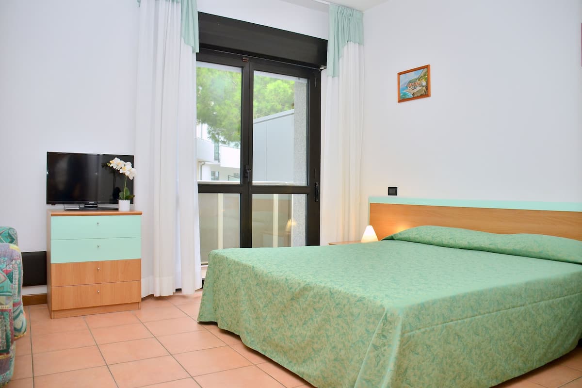Ein Schlafzimmer mit einem Bett, einem Fernseher auf einer Holzkommode und einem Fenster mit Vorhängen.