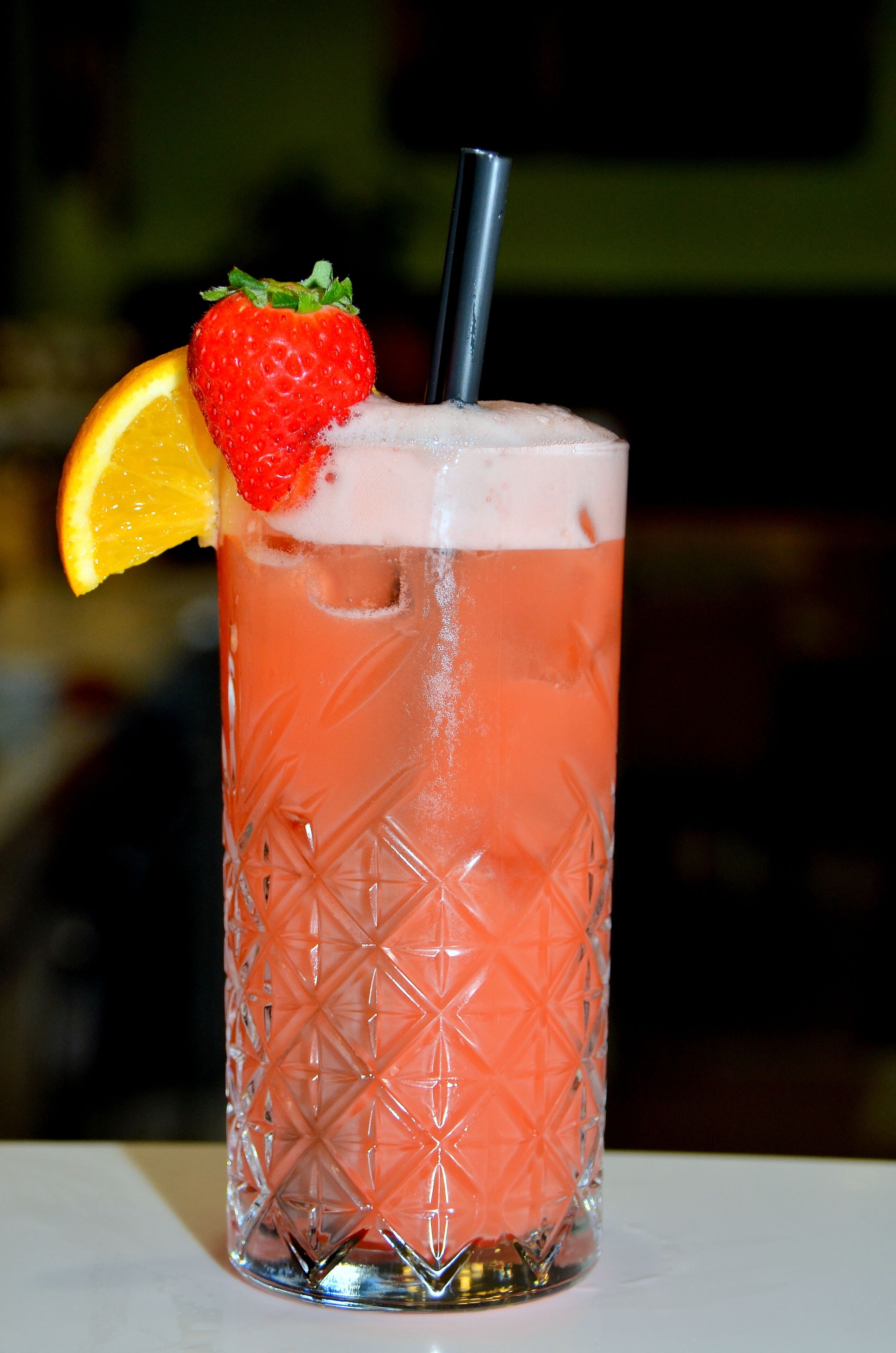 Un grand verre à motifs, rempli d’une boisson rosée, agrémenté d’une fraise et d’une tranche d’orange, et muni d’une paille.