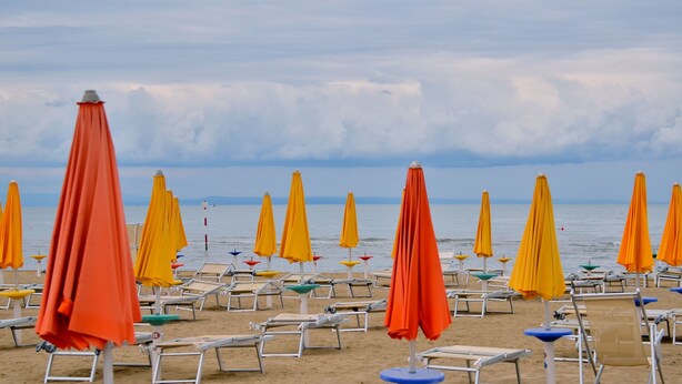 Ein Strand mit Reihen von Liegestühlen und Sonnenschirmen, einige mit orangefarbenen und gelben Markisen, unter einem bewölkten Himmel.