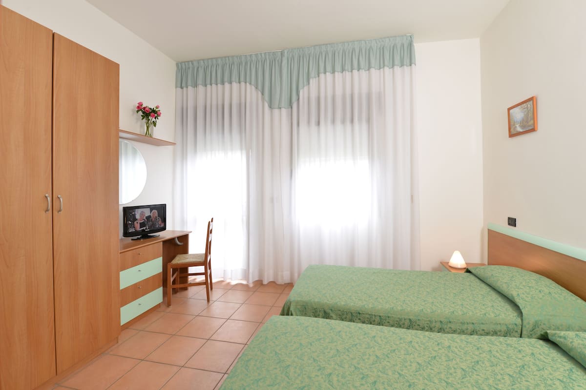 Ein Schlafzimmer mit zwei Betten, einem Schreibtisch, einem Stuhl, einem Kleiderschrank und einem Fenster mit Vorhängen.