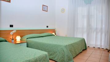 Quarto de hotel com duas camas, uma mesa de cabeceira com abajur, relógio na parede e janela com cortinas finas.