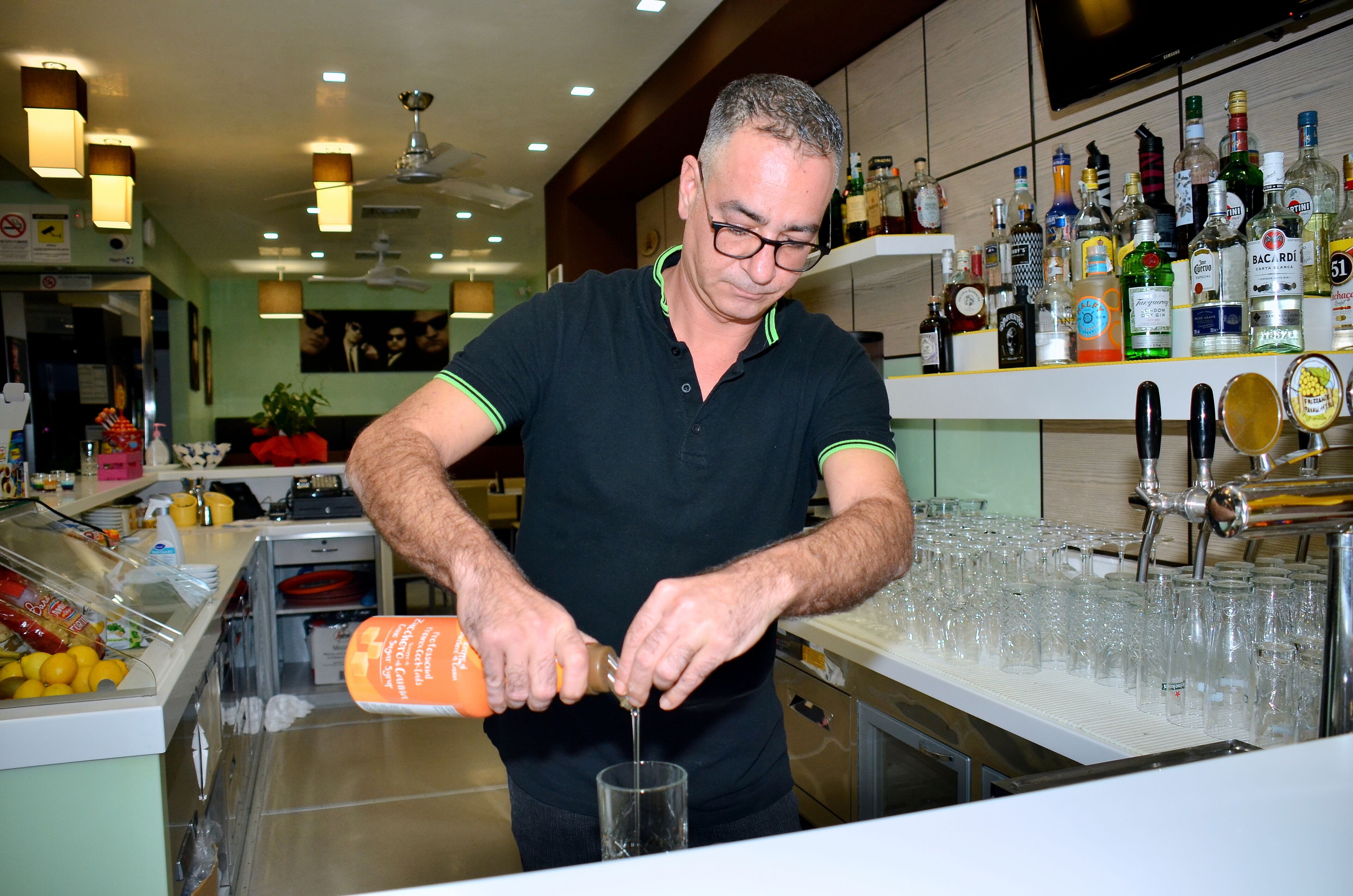 Zona de bar con un camarero sirviendo una bebida, una variedad de botellas en estantes y una barra con vasos.