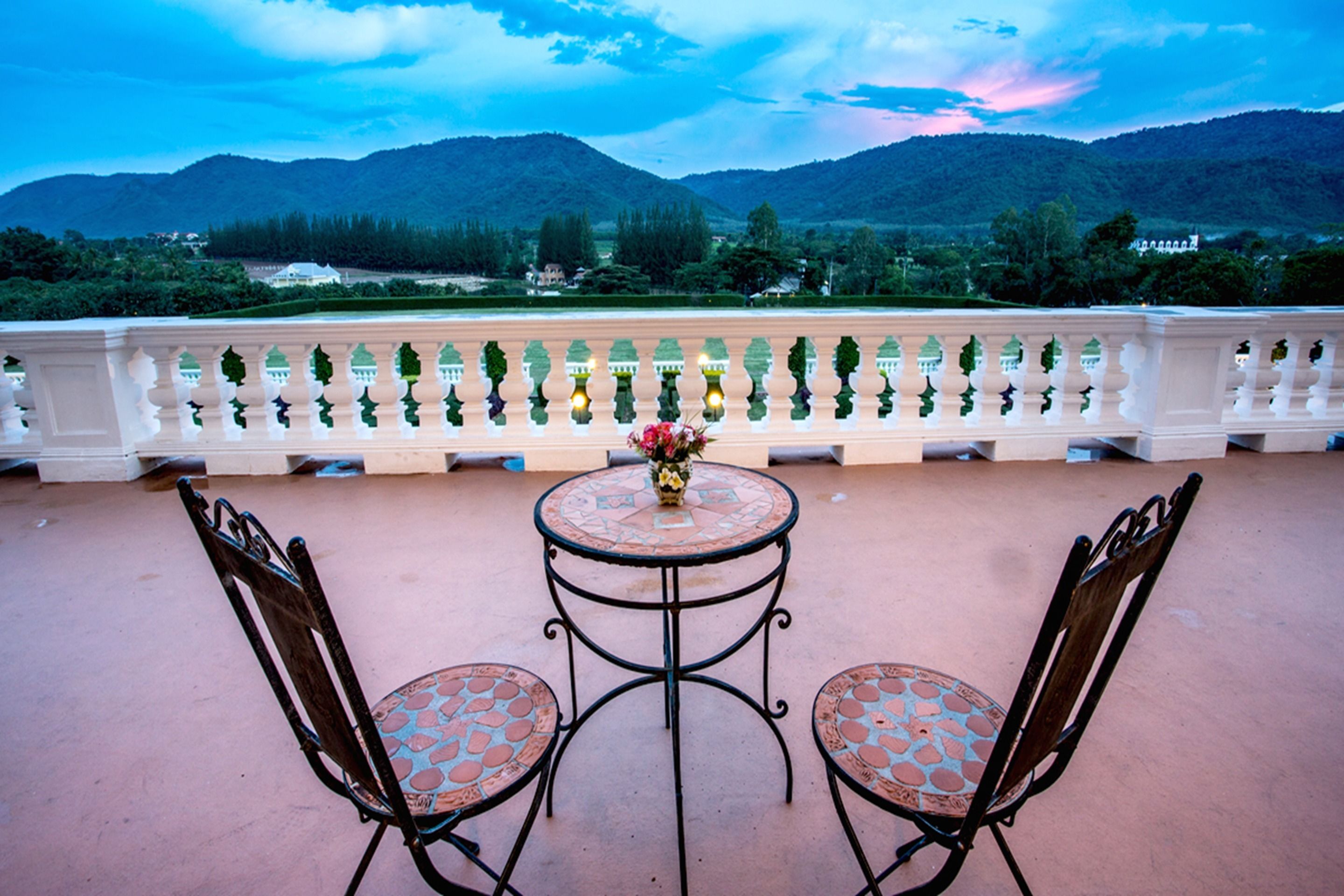 A terrace with a table and two chairs, a white balustrade, and a view of mountains.