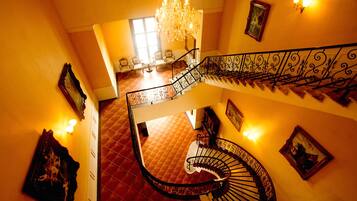 Una imponente escalera con barandillas ornamentadas y un candelabro, que conduce a una habitación con piso de baldosas rojas y un amplio ventanal.