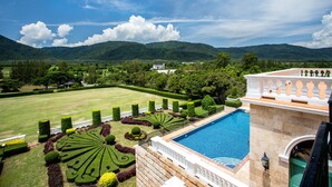 Une piscine entourée d’un jardin à la disposition géométrique, offrant une vue sur un paysage montagneux, et un bâtiment à l’architecture classique.