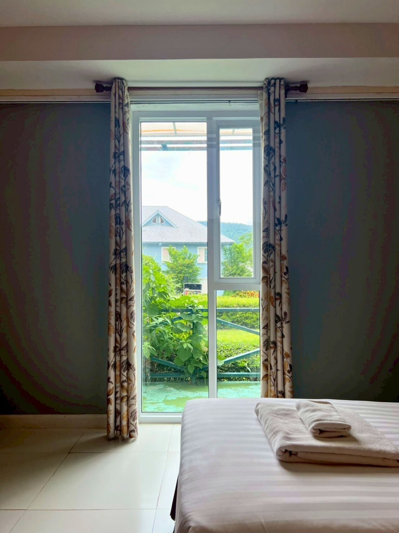 Une chambre avec une grande fenêtre, des rideaux, un lit avec une serviette pliée et une vue sur la verdure à l’extérieur.