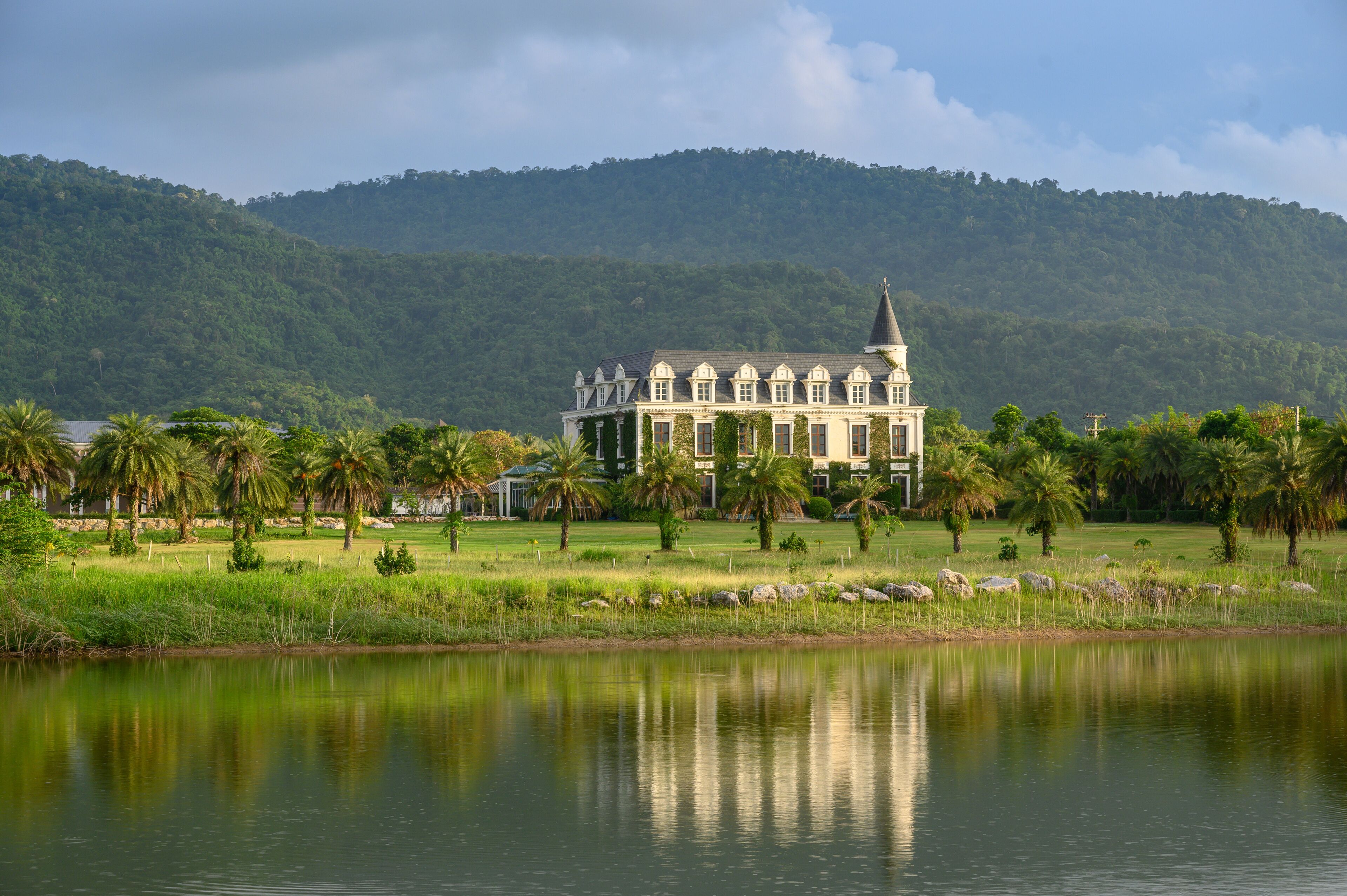 Um edifício grande e branco com uma torre, rodeado por uma paisagem verdejante e um corpo de água calmo.
