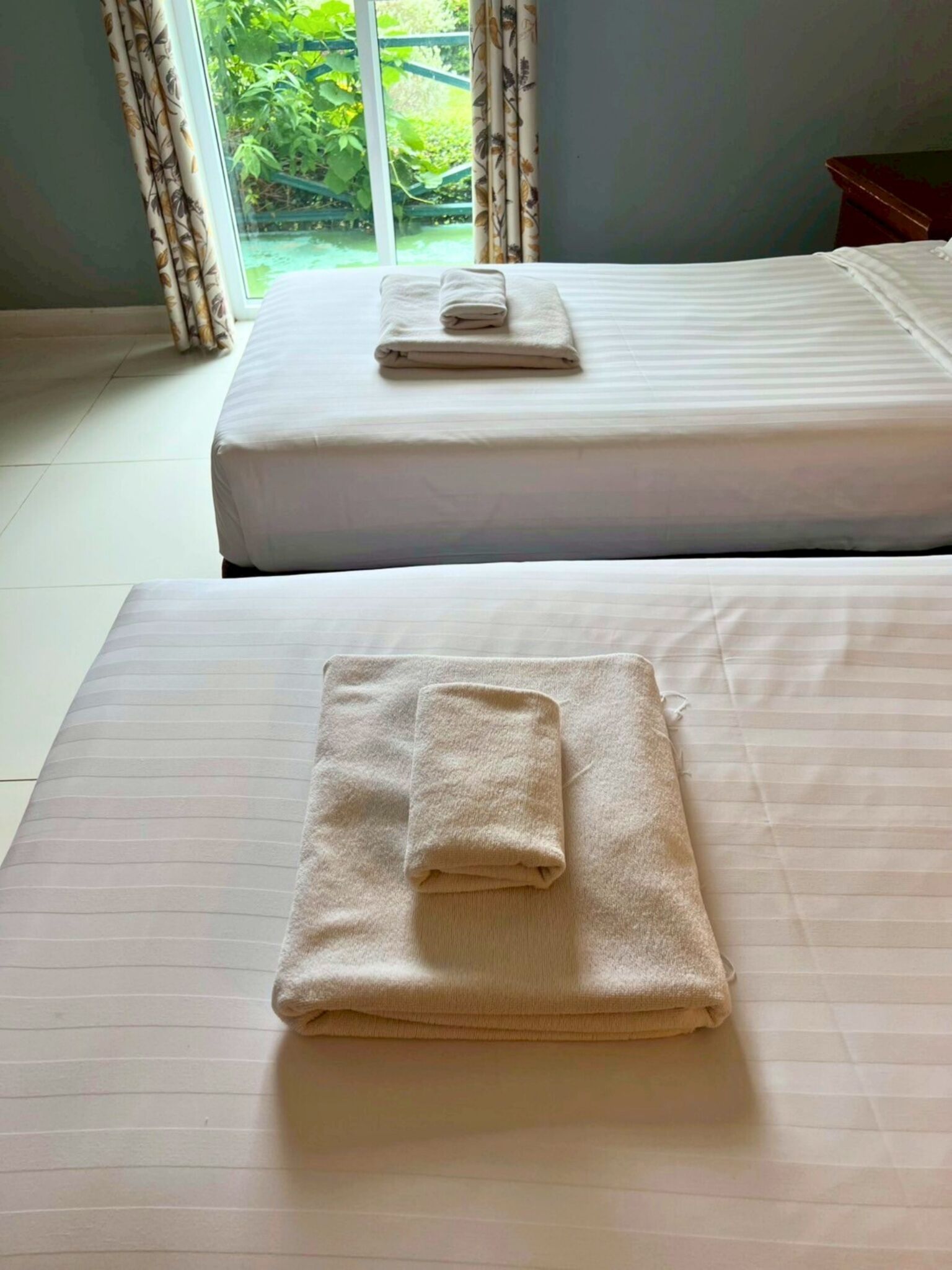 Two beds with neatly folded towels, a window with curtains, and a view of greenery outside.