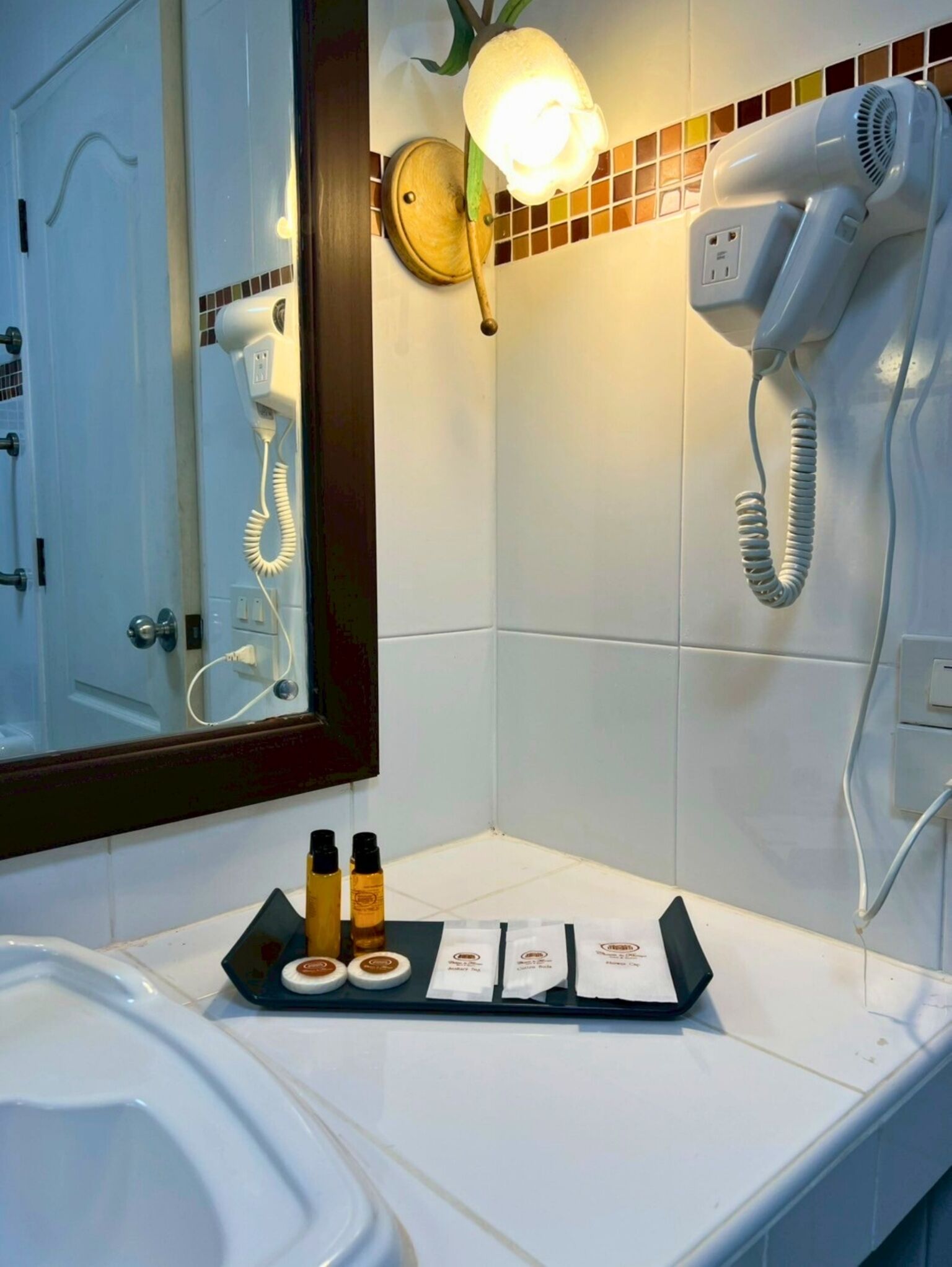 A bathroom with a hairdryer mounted on the wall, a mirror, and a tray with toiletries.