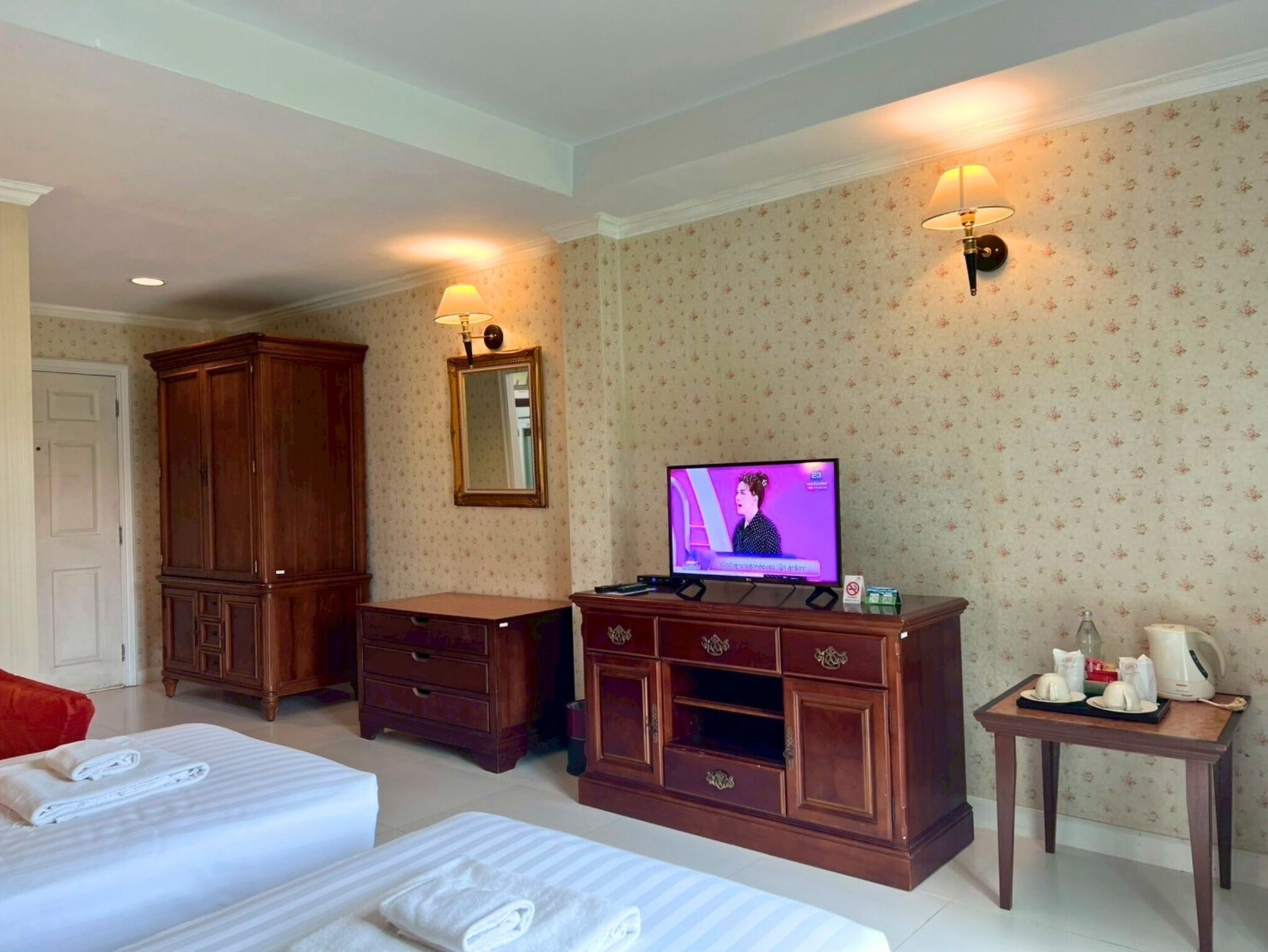 A hotel room with two beds, a wooden dresser, a television, and a small table with a kettle and cups.