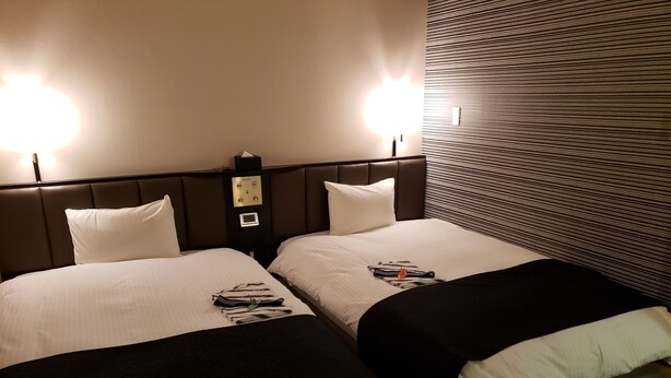 A hotel room with two beds, each with a headboard, a bedside table, and a lamp. - APA Hotel Owariichinomiya Station (Ichinomiya)