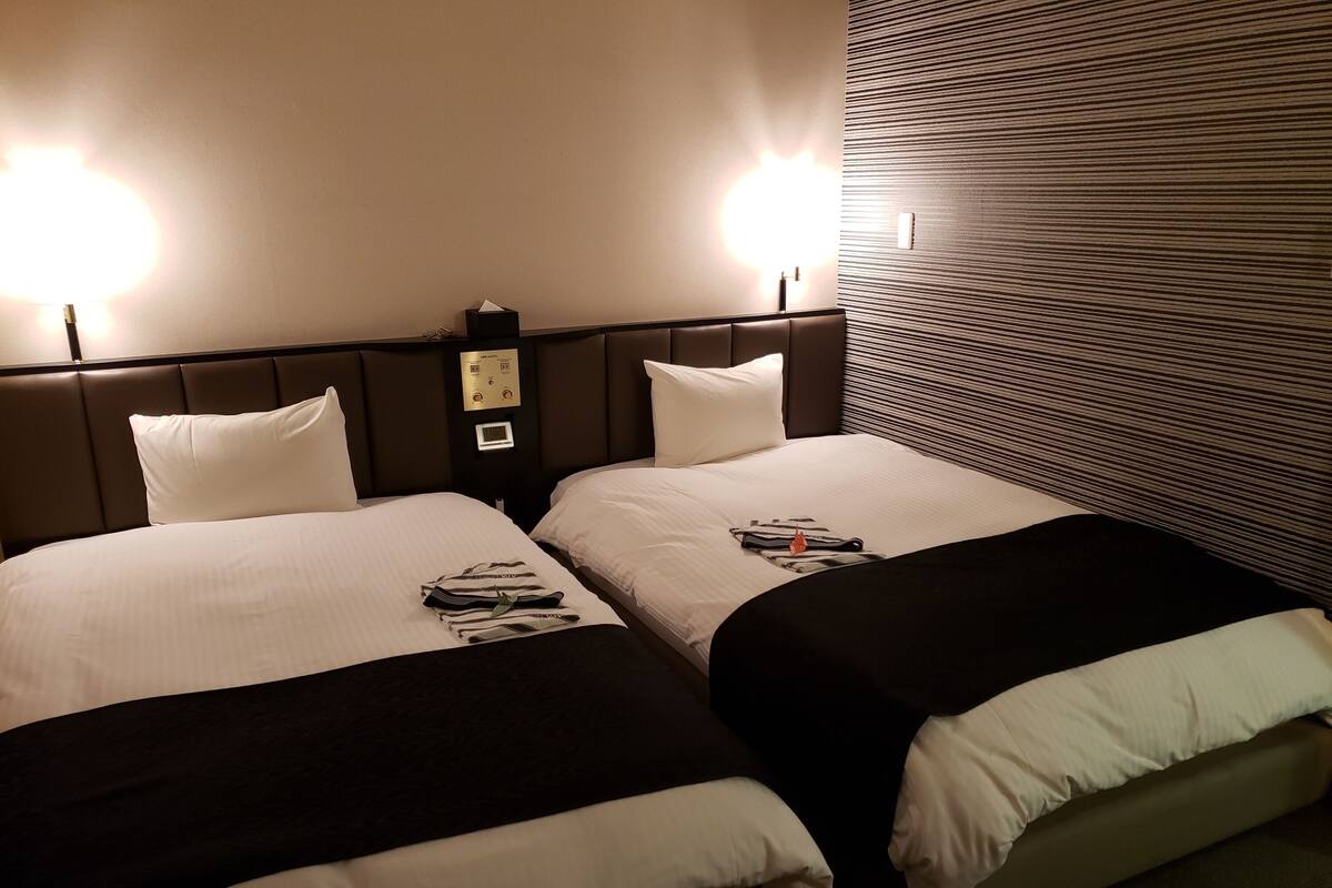 A hotel room with two beds, each with a headboard, a bedside table, and a lamp.