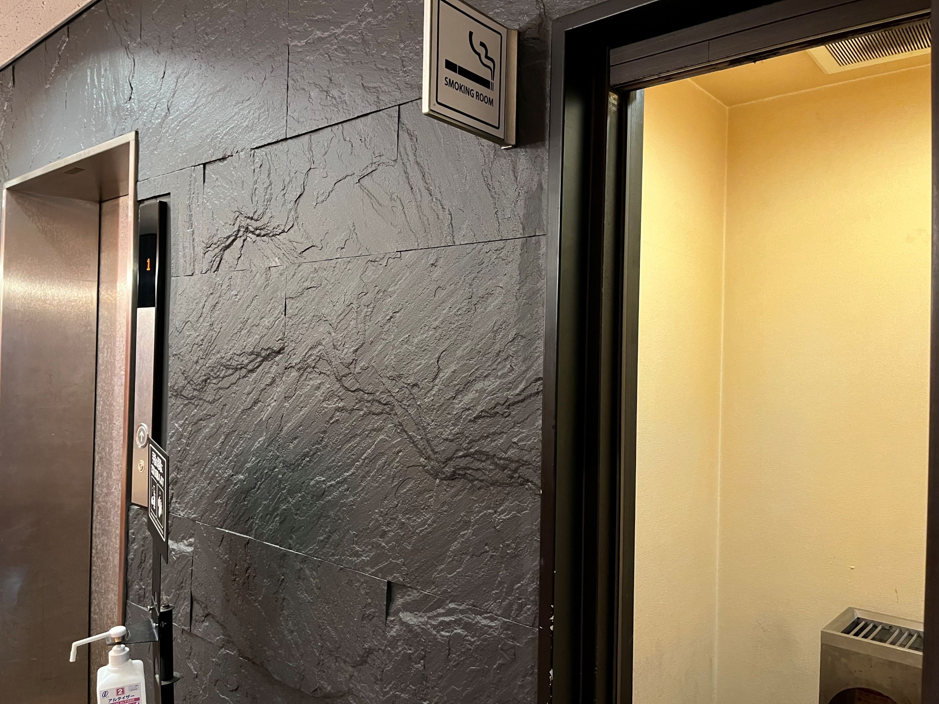 Pared texturizada con un cartel de 'Smoking Room', dispensador de gel hidroalcohólico y puerta de ascensor.