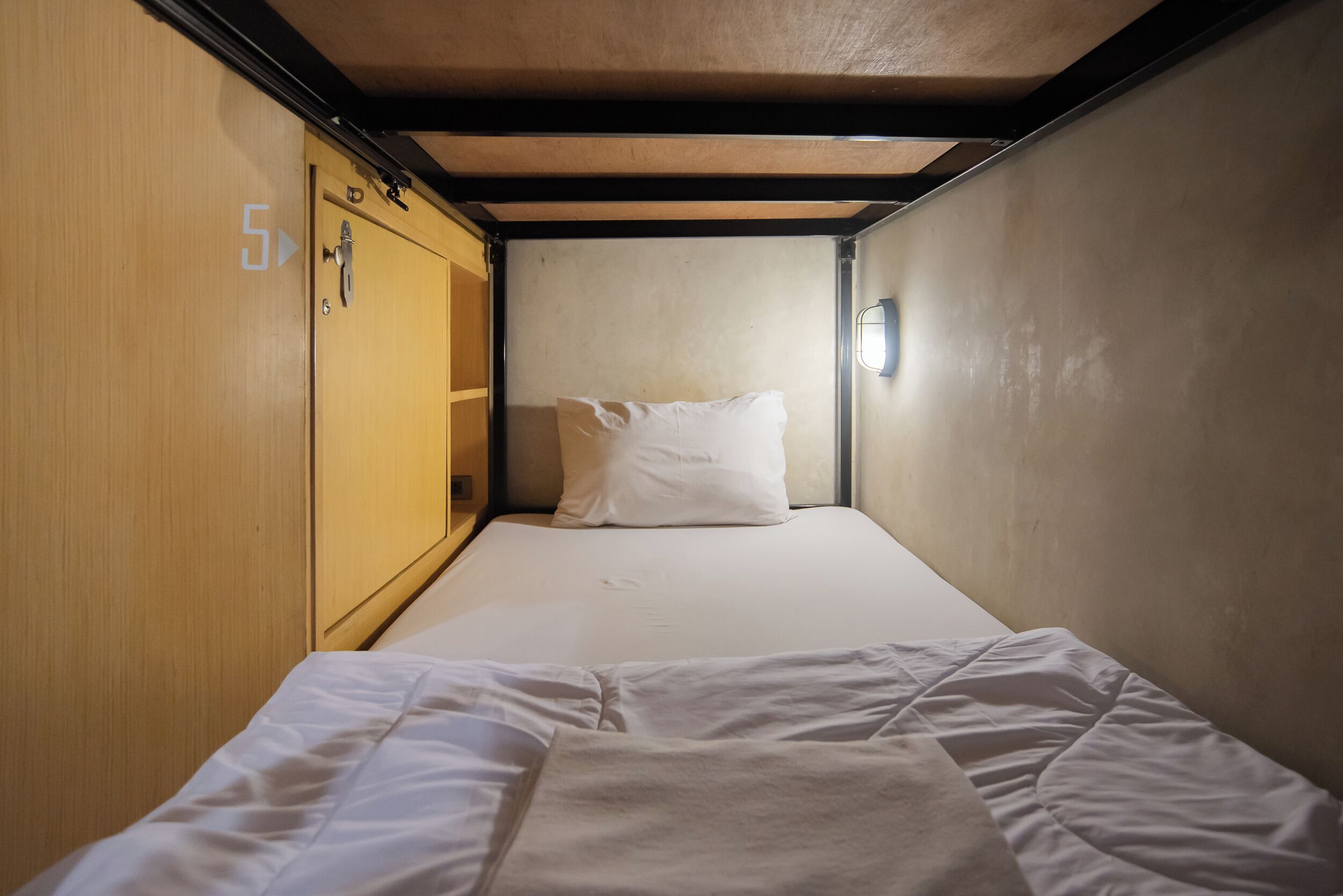 A narrow, single-bed room with a wooden wardrobe on one side and a wall-mounted light fixture.