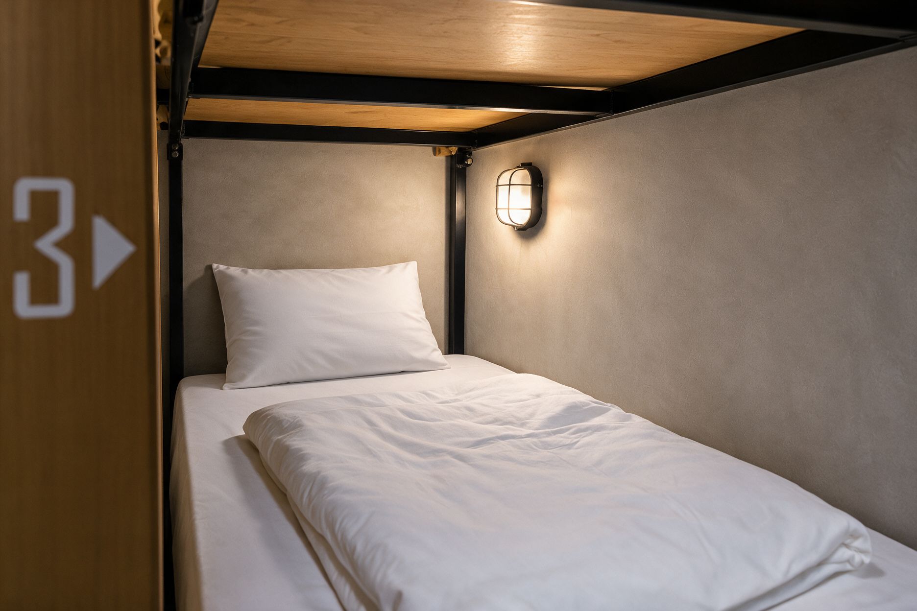 Un lit simple avec un oreiller et un luminaire fixé au mur, dans une pièce dont le plafond est en bois et dont la porte porte un numéro.