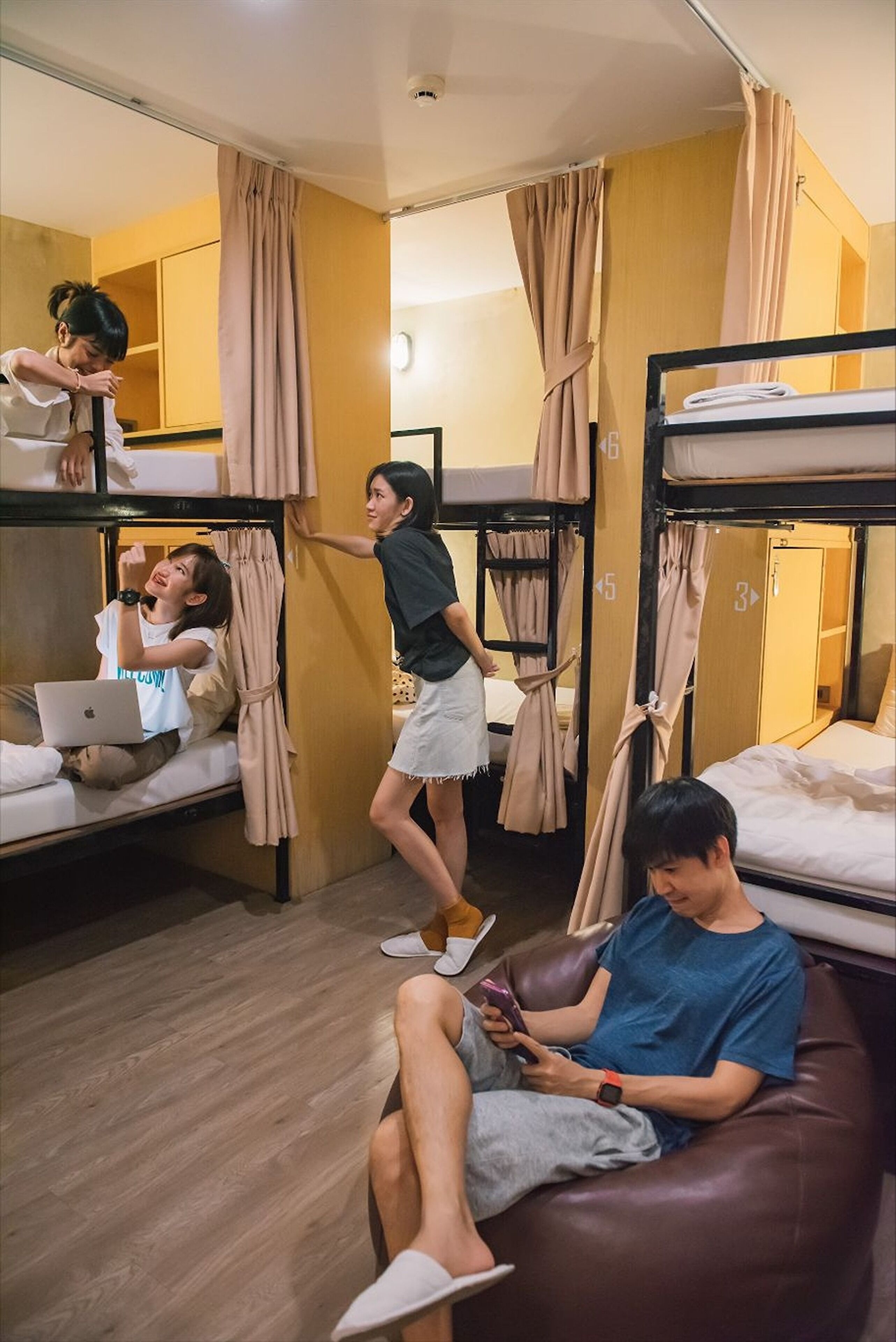 Ein Schlafsaalzimmer mit Etagenbetten, ein Laptop auf einem Bett und eine Person, die ein Smartphone benutzt.