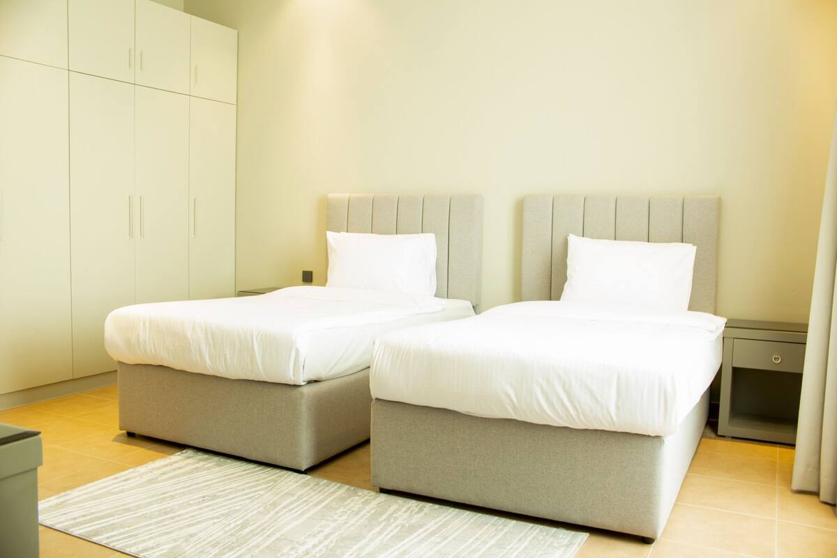 A hotel room with two single beds, a grey headboard, a small bedside table, and a wardrobe.