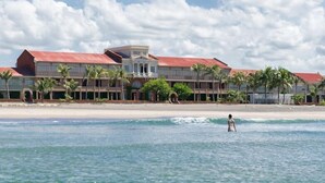 Een hotel aan het strand met een groot gebouw, palmbomen en een persoon in het water.