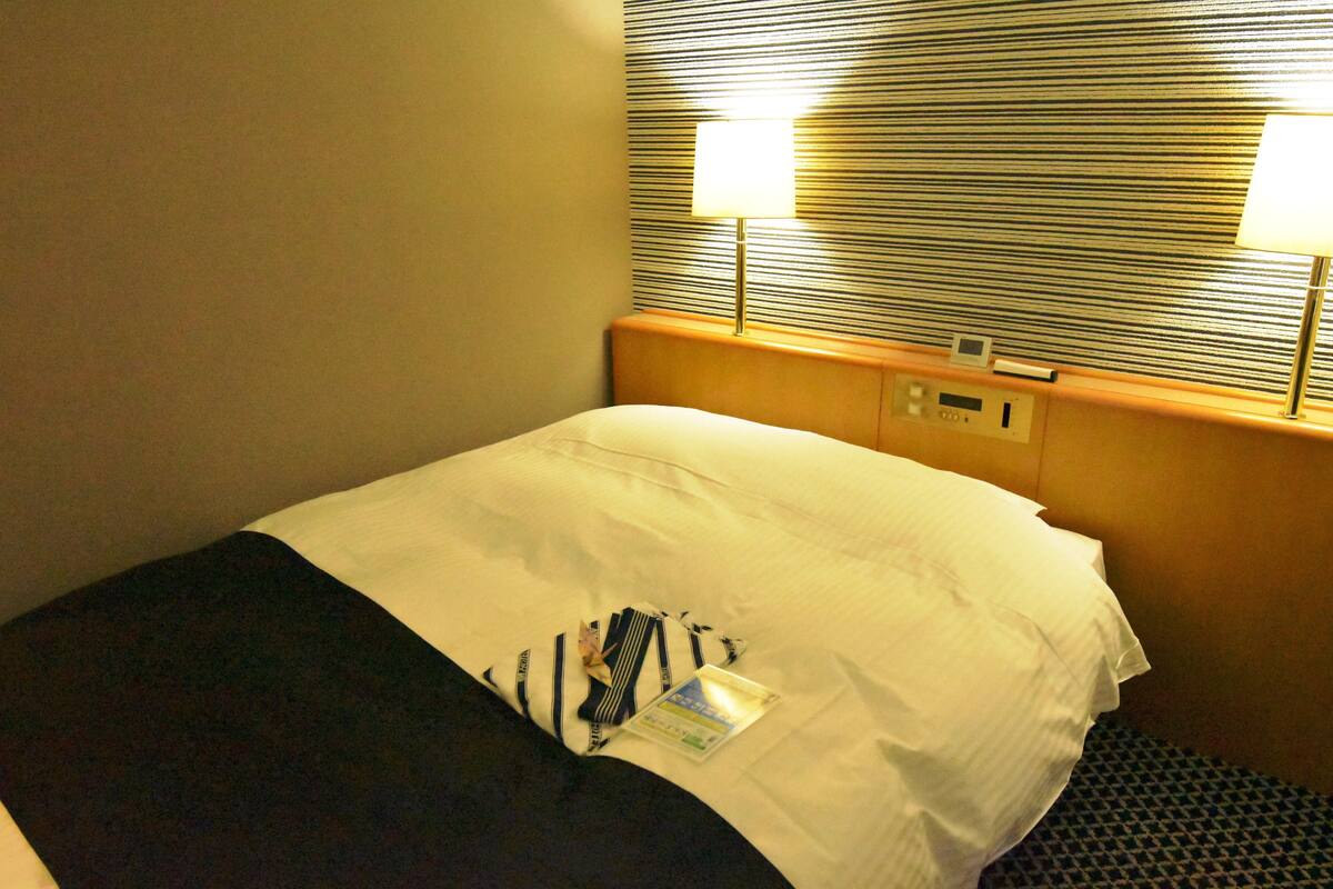 Ein Hotelzimmer mit Bett, Kopfstütze, zwei Nachttischlampen und einem Fenster mit Jalousien.