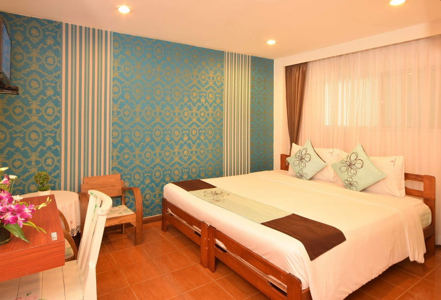 A hotel room with a large bed, a wooden chair, a small table with a vase of flowers, and a patterned wall.