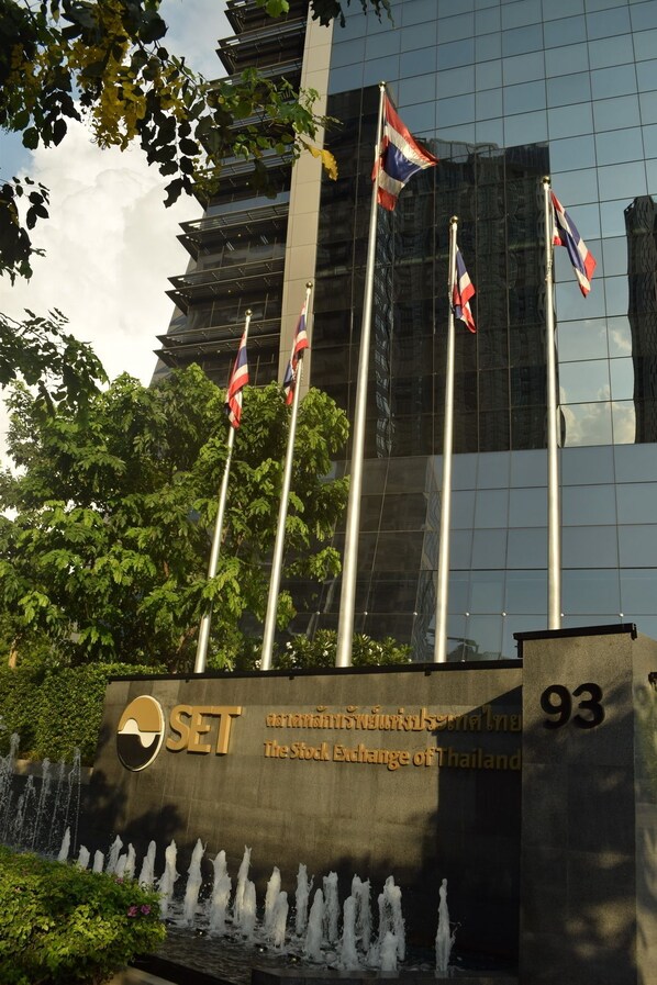 The exterior of the Stock Exchange of Thailand with flags, a water feature, and a modern glass building in the background. - Blissotel Ratchada (Bangkok)