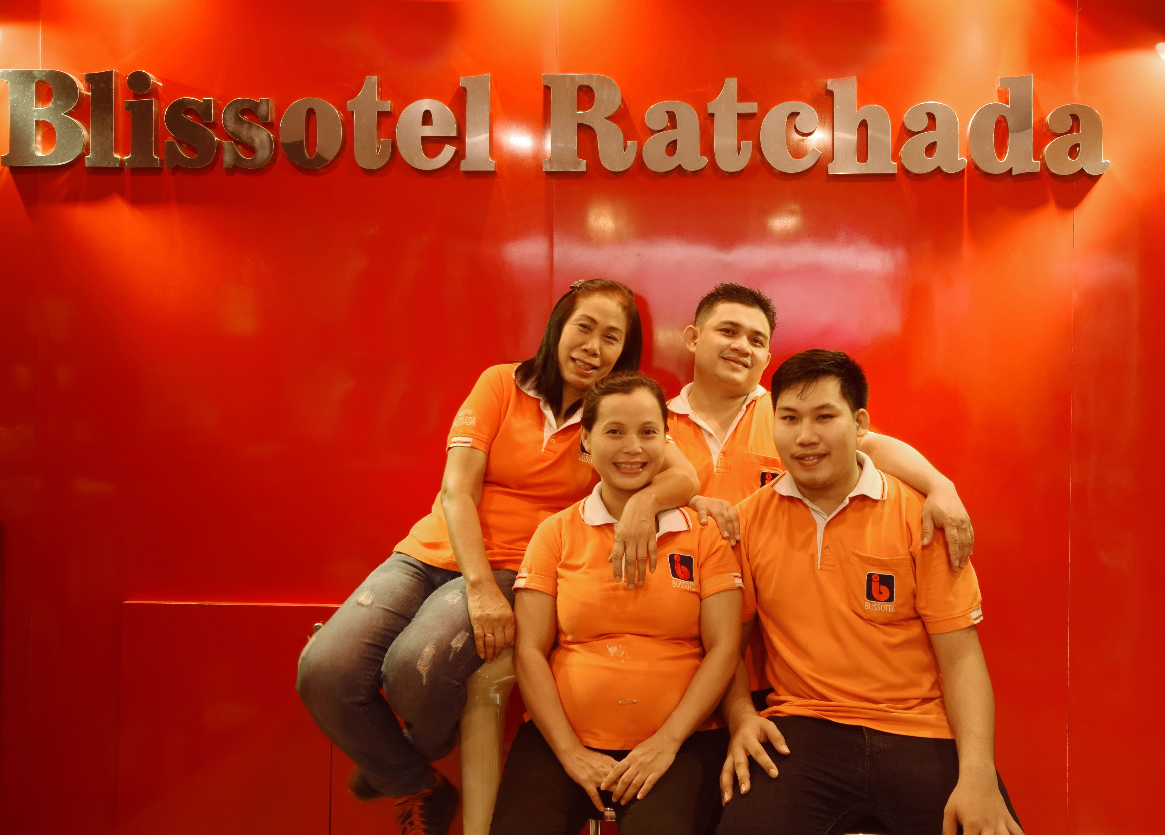A group of people in orange uniforms posing in front of a red wall with the text "Blissotel Ratchada".