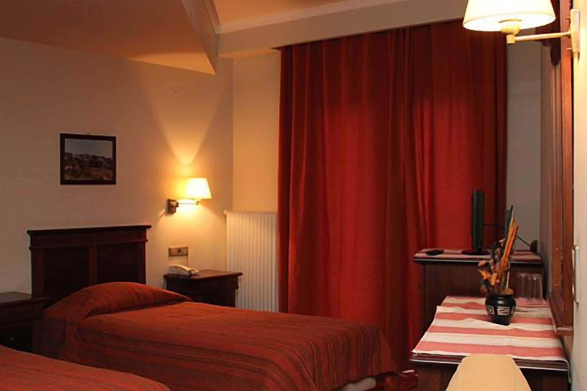A hotel room with two beds, a table with a vase, a lamp, and a red curtain.