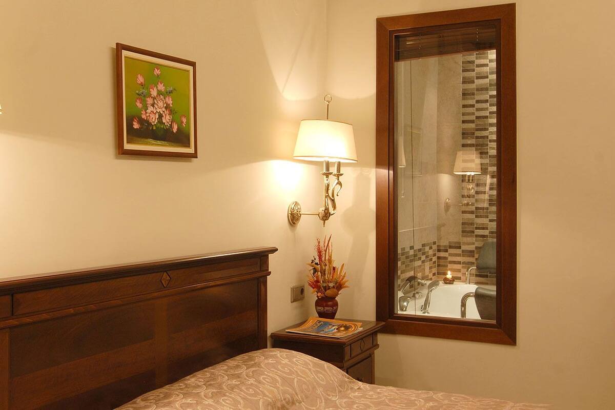 Et hotelværelse med sengegavl i træ, et indrammet blomsterbillede, en natlampe, en vase med blomster og et stort spejl der afspejler et badeværelse med flisebeklædt bagvæg.