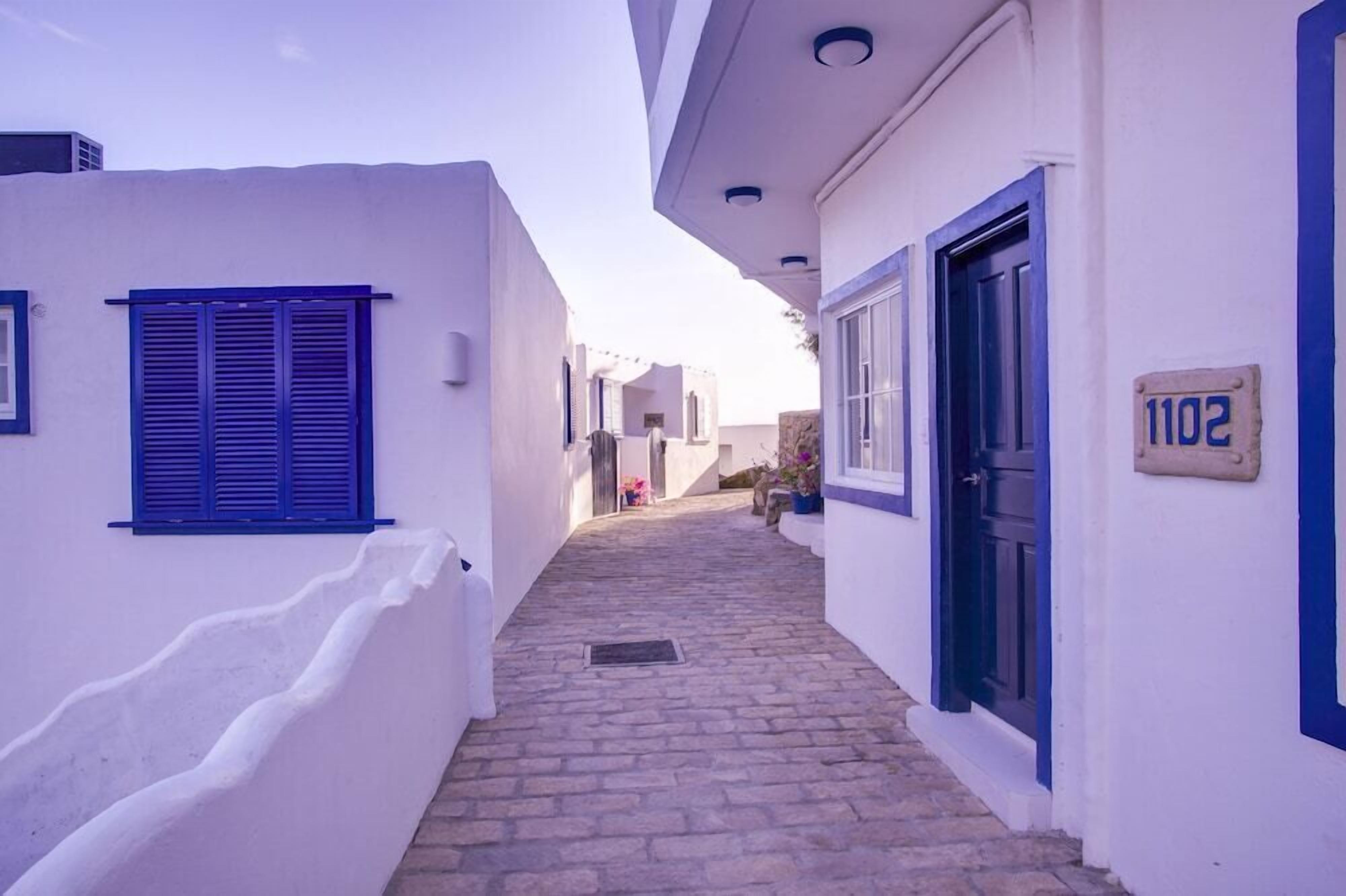 A narrow cobblestone alleyway between white buildings with blue shutters and doors, leading to a street.