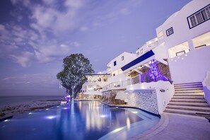 Vista a la piscina de un edificio blanco con balcones, un árbol y vistas al mar.