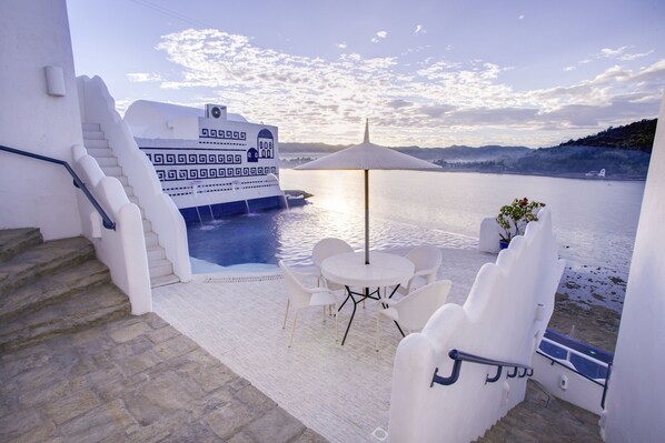 Un balcón con zona de estar exterior blanca, vista a un cuerpo de agua y un barco blanco al fondo.
