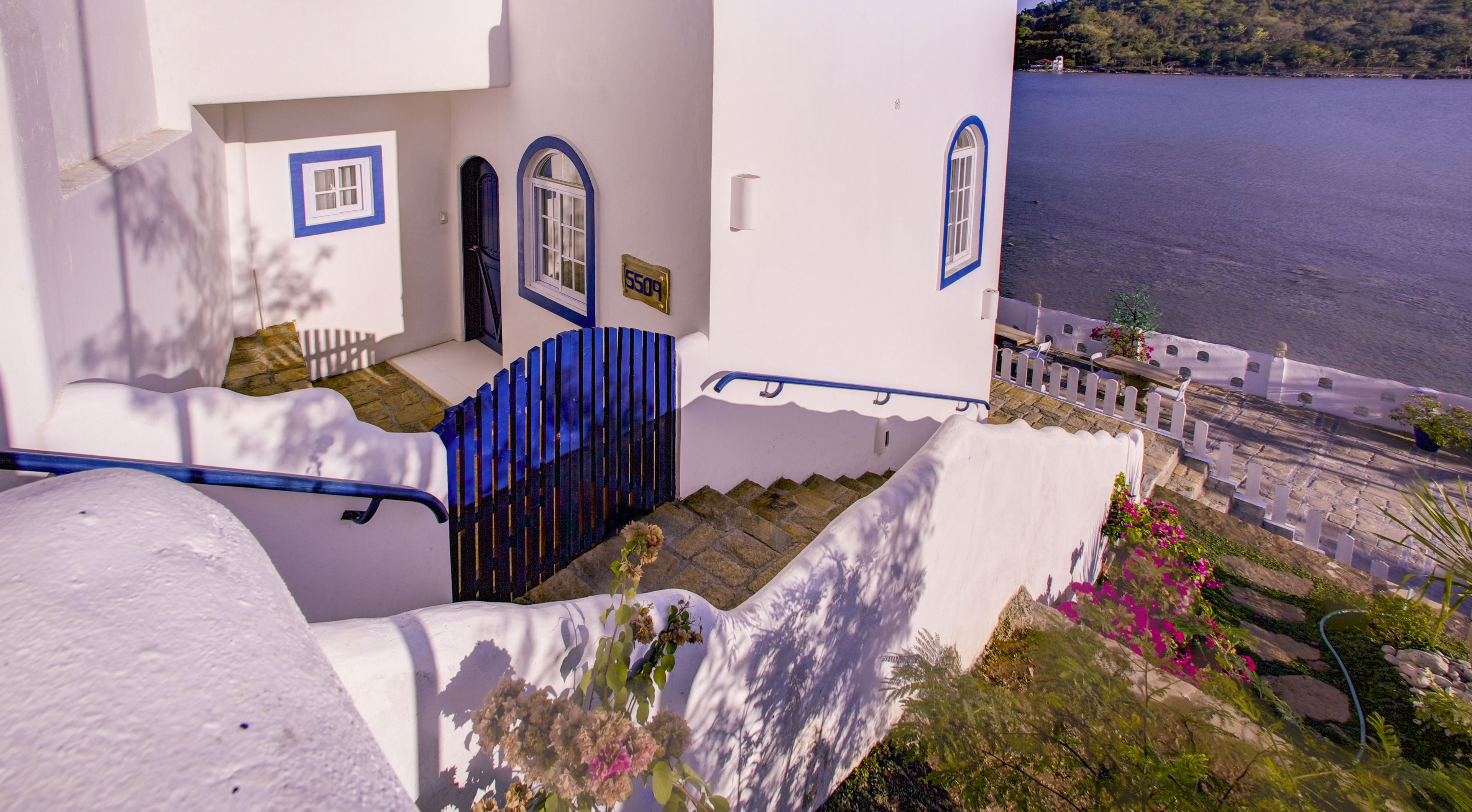 Un edificio blanco con detalles azules, una escalera que lleva a una puerta y vista a un cuerpo de agua.