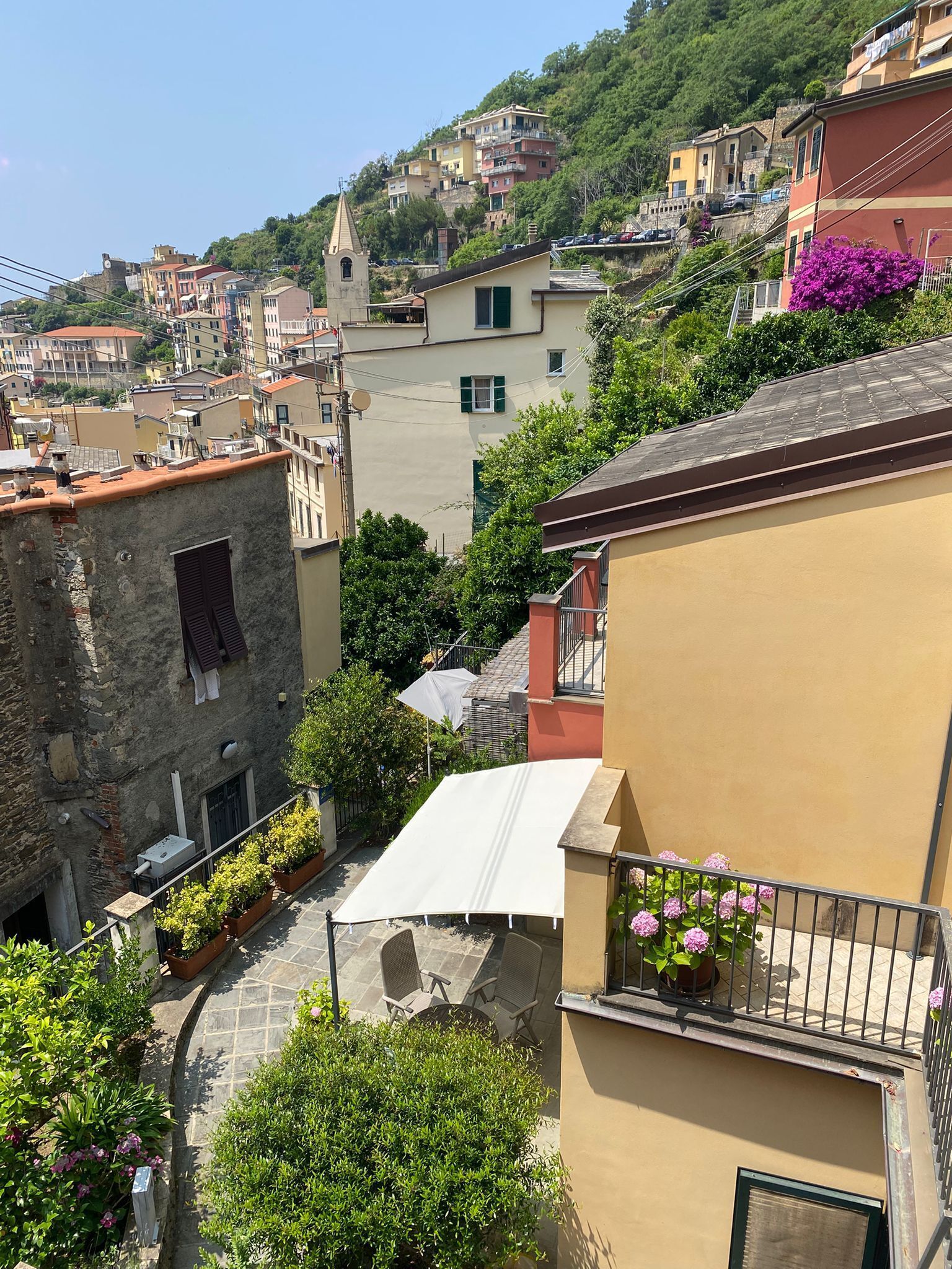 A narrow street with buildings on both sides, a small courtyard with a white awning, and a view of a hillside with colorful houses.