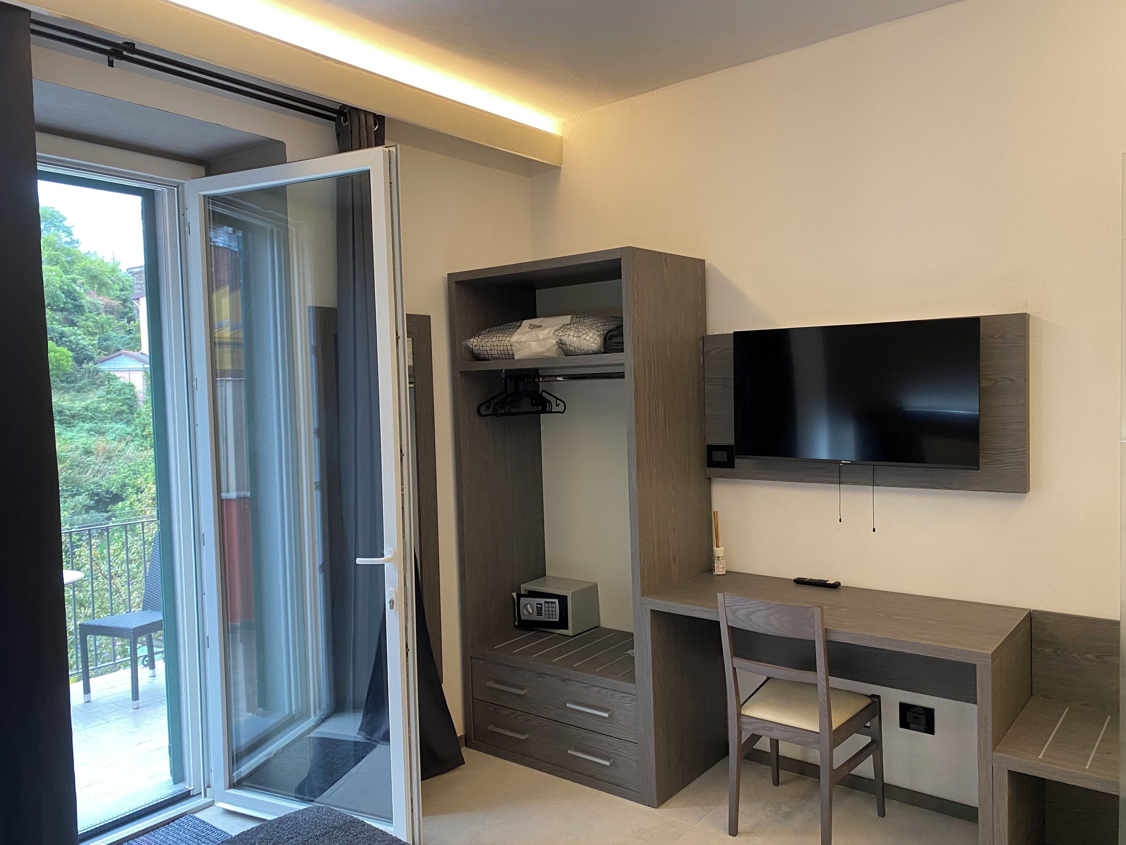 Une chambre moderne dotée d’une porte-fenêtre coulissante donnant sur un balcon, d’une armoire intégrée, d’un bureau avec une chaise et d’un téléviseur fixé au mur.