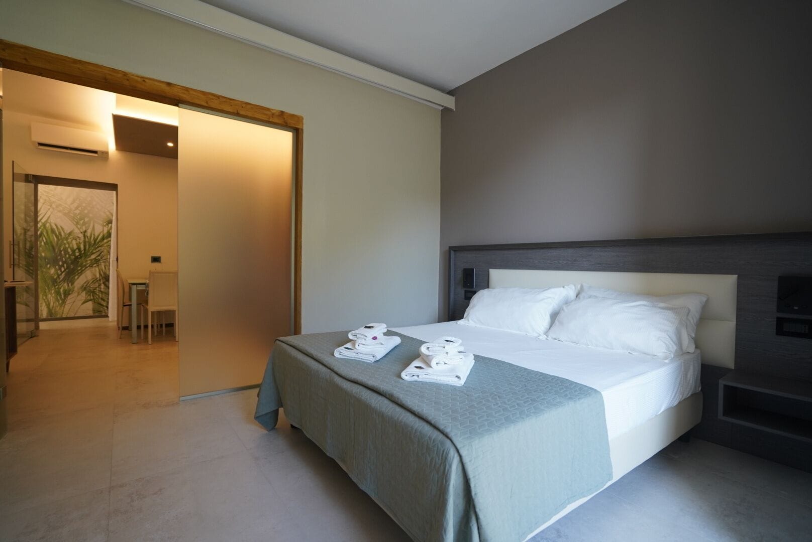 A modern hotel room with a large bed, a bedside table, and a view of a dining area.