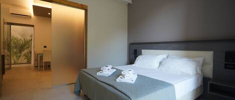 A modern hotel room with a large bed, a bedside table, and a view of a dining area.
