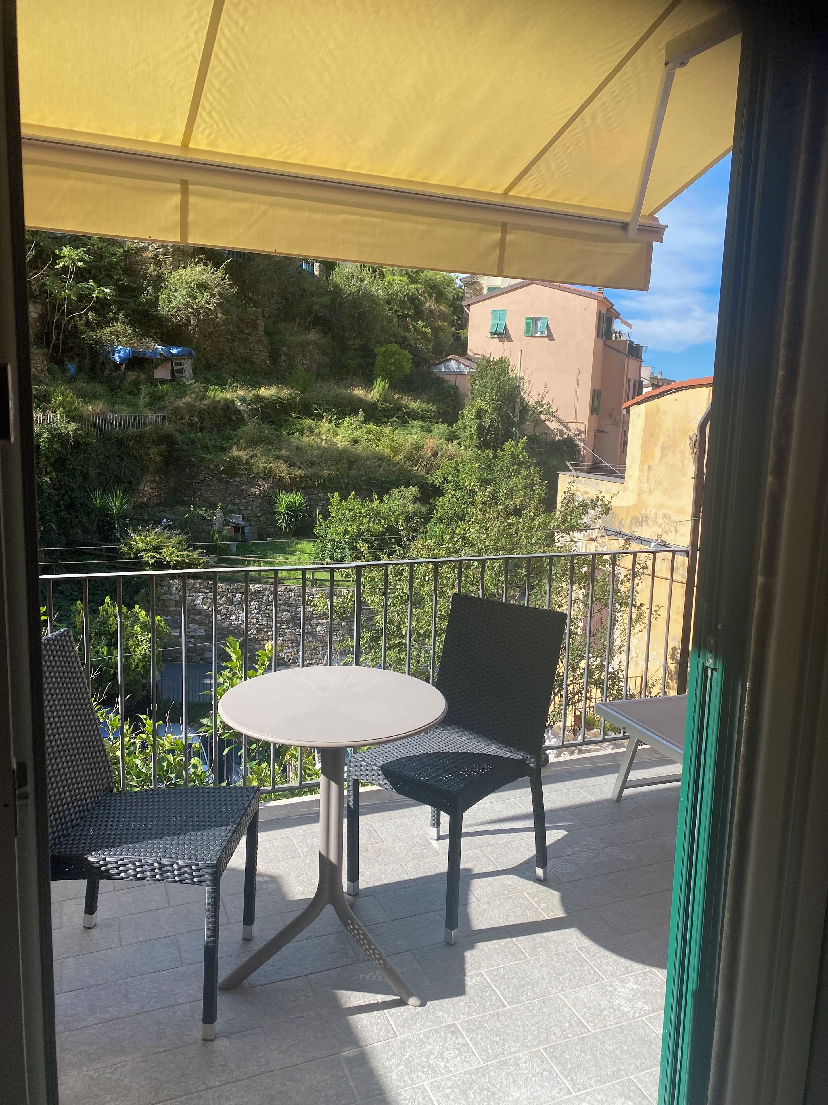 A balcony with a table and two chairs, a yellow awning, and a view of a hillside with buildings.