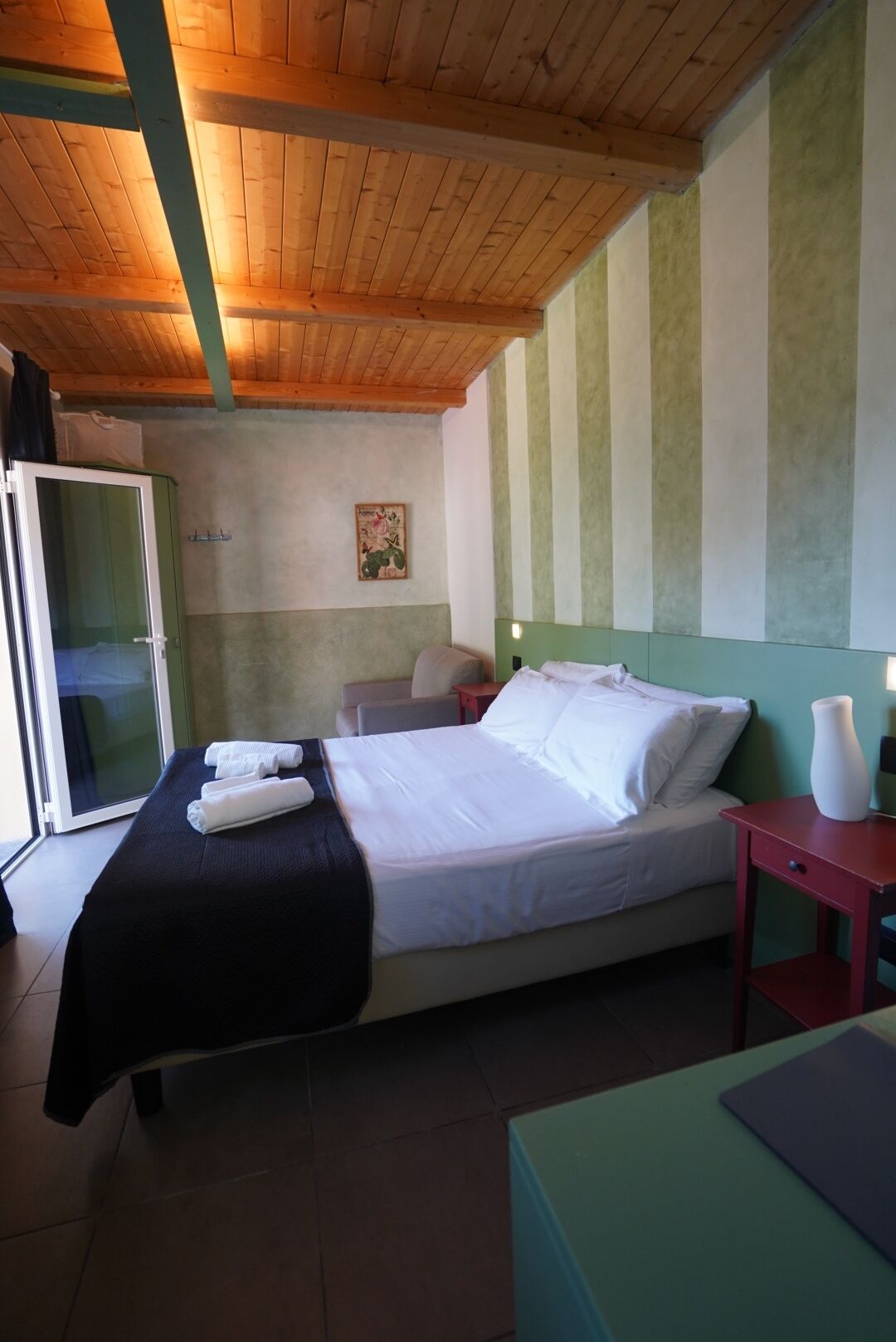 A single-bed room with a wooden ceiling, a green-striped wall, a bedside table with a vase, and a sliding door to the left.