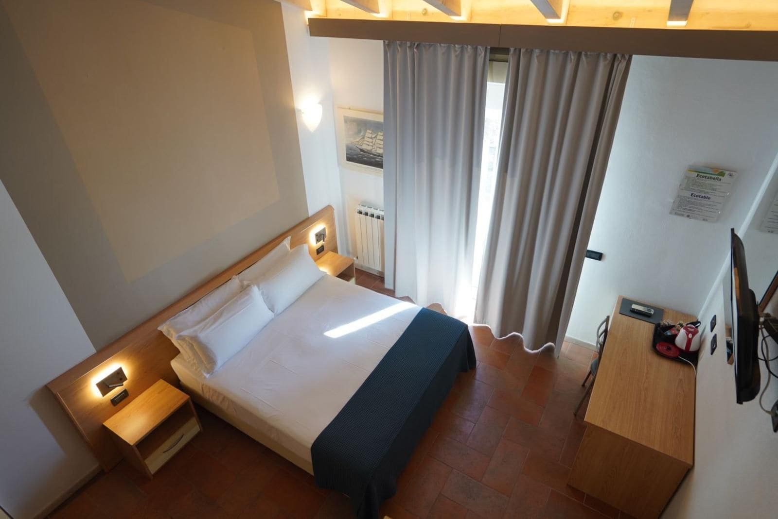 Habitación de hotel con cama, mesitas de noche, escritorio, silla, televisor y ventana con cortinas.