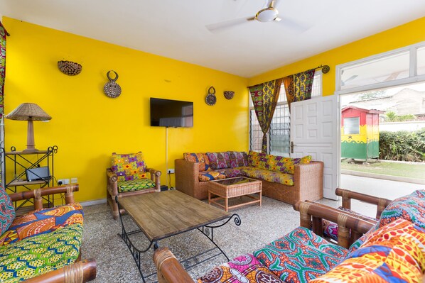 A bright yellow room with a sofa, a wooden coffee table, a TV mounted on the wall, and a ceiling fan. - Agoo Hostel (Accra)