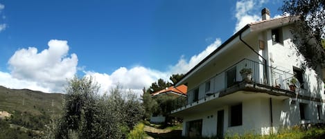 A two-story house with a balcony, surrounded by trees and a mountainous landscape.