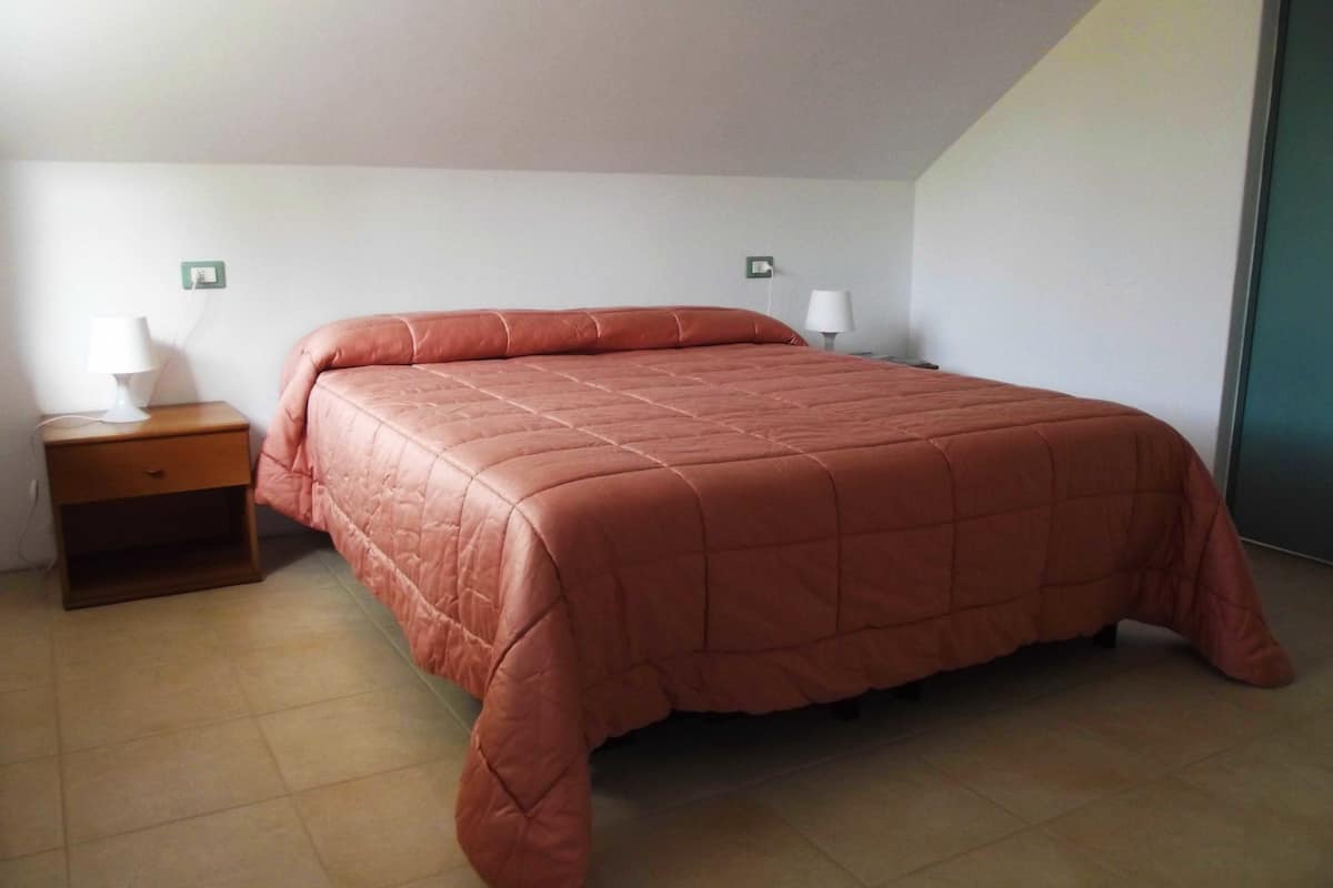 A bedroom with a large bed, a nightstand, a lamp, and a wall socket.