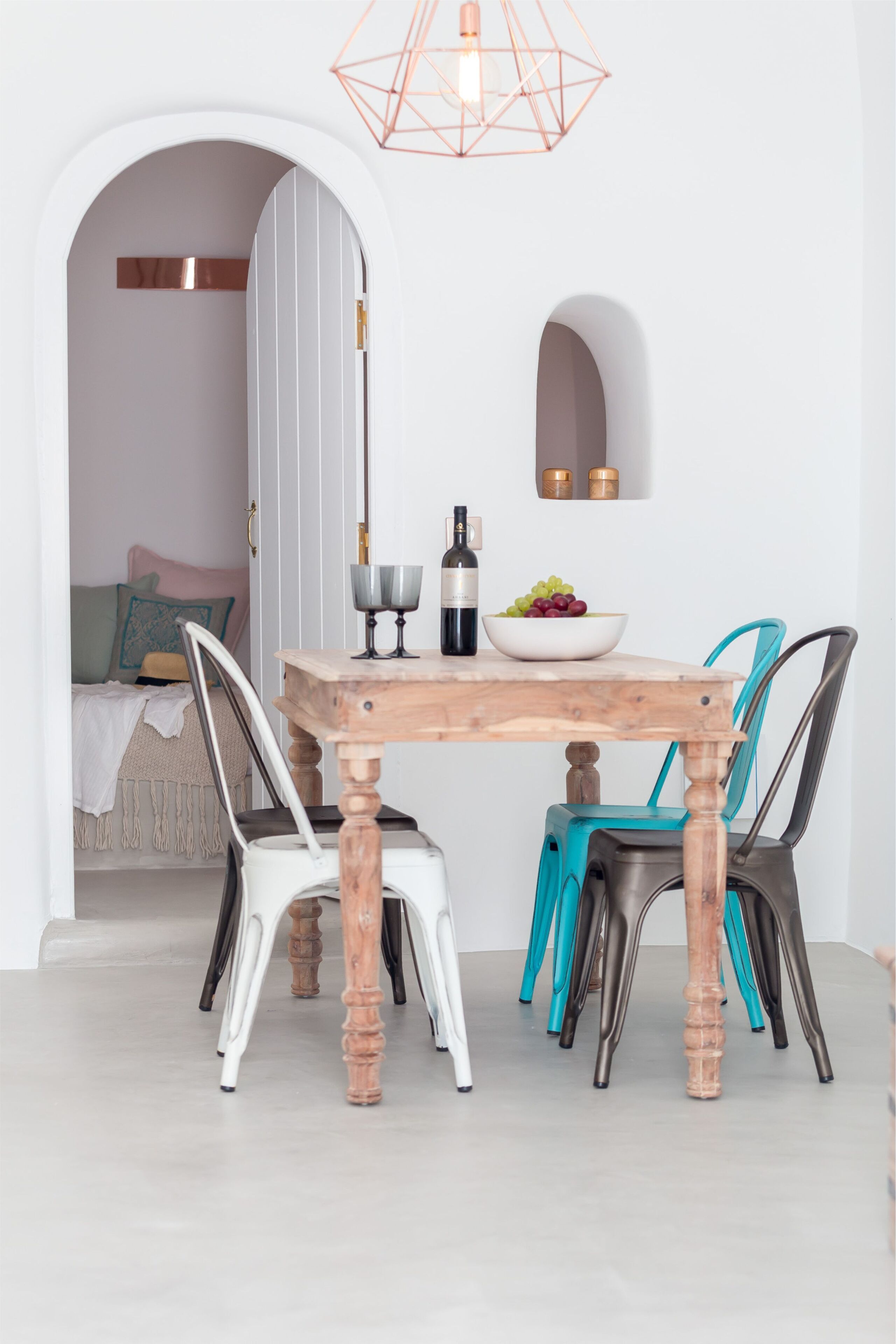 A dining area with a wooden table, four chairs, a bowl of fruit, a wine bottle, and a geometric pendant light.