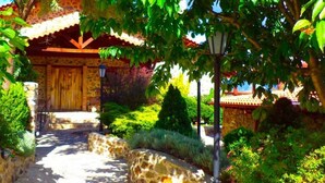A stone pathway leading to a rustic wooden building with a tiled roof, surrounded by lush greenery and flowering plants. - Loggas Hotel (Kastoria)