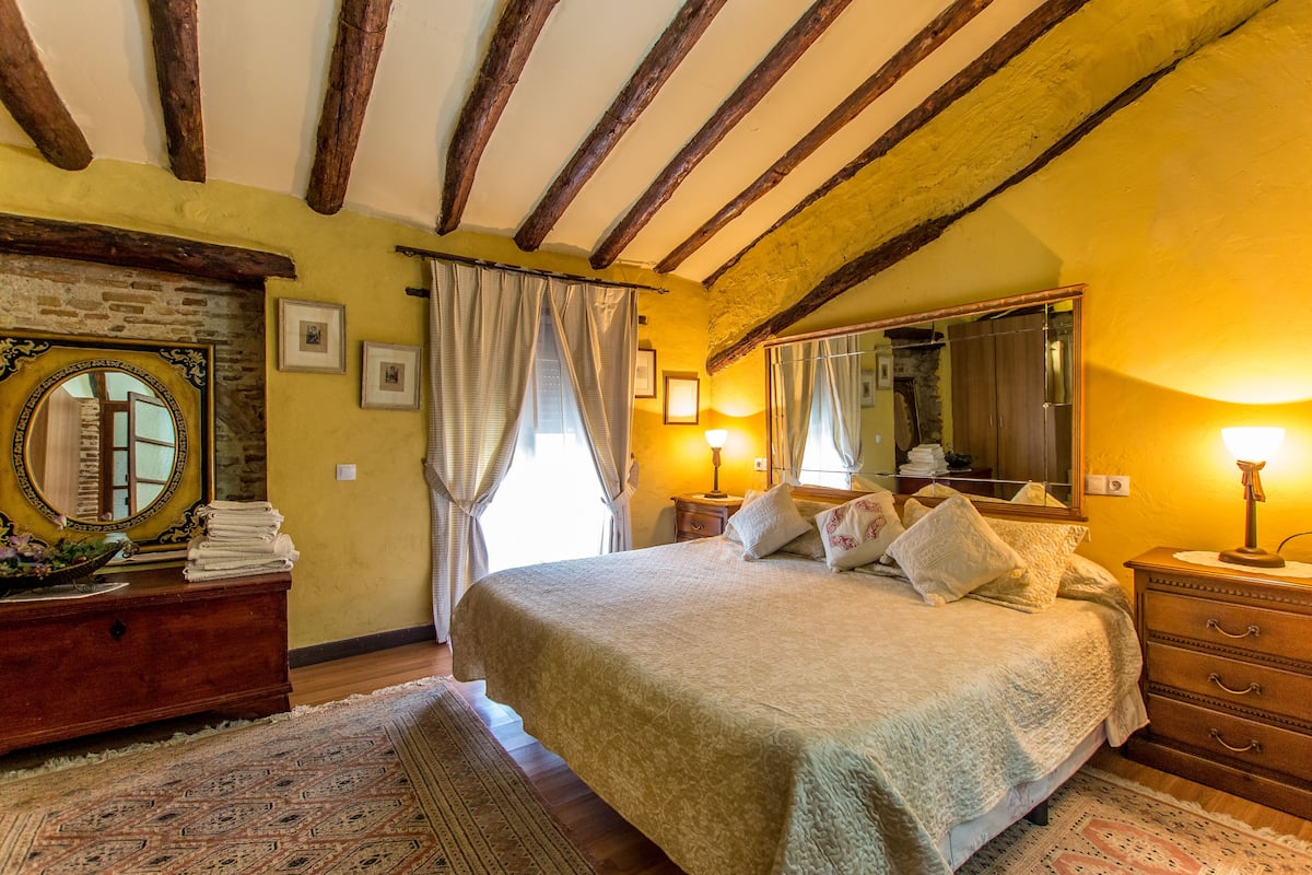A bedroom with a large bed, wooden beams on the ceiling, a dresser, and a mirror.