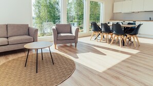 Una sala de estar moderna con sofá, sillón y una mesa redonda, un comedor con sillas y ventanas amplias que ofrecen vistas a árboles.