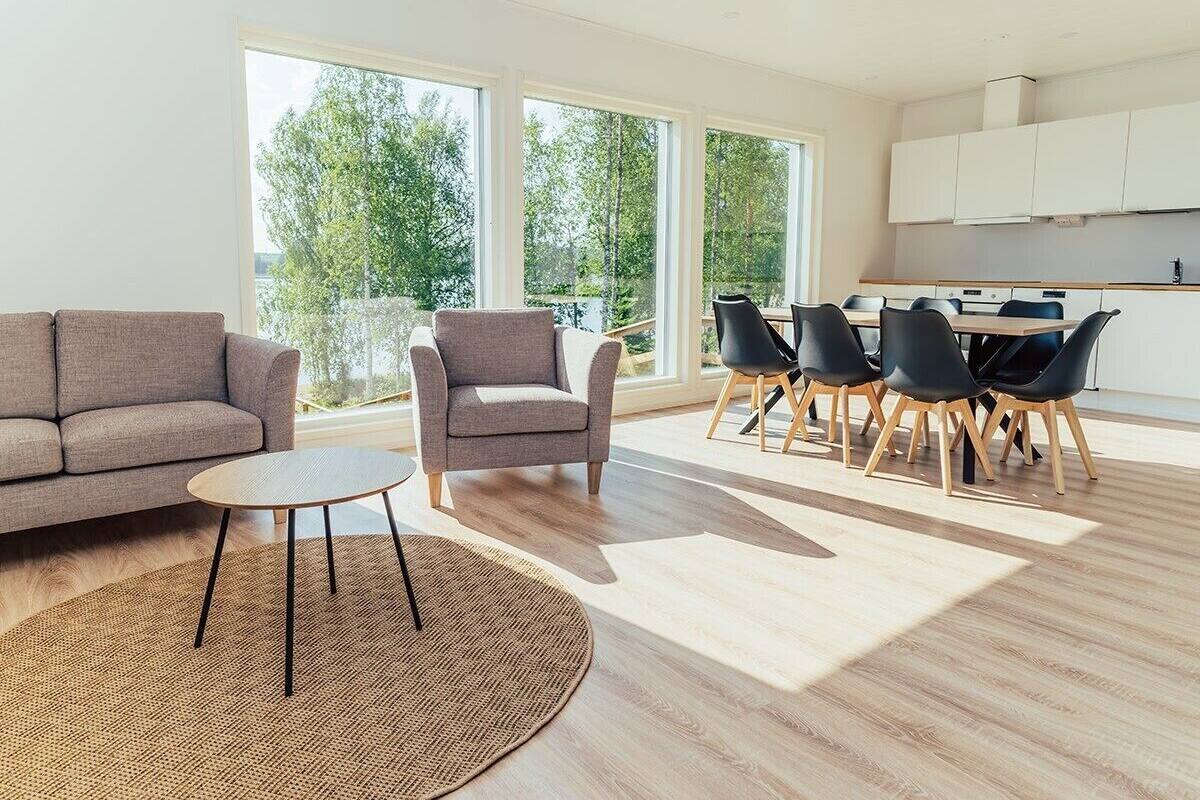 Ein modernes Wohnzimmer mit Sofa, Sessel und rundem Tisch, ein Essbereich mit Stühlen und großen Fenstern mit Blick auf Bäume.