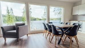Un comedor moderno con una gran mesa y sillas, ventanas amplias con vistas a un lago y un bosque, y una cocina con armarios blancos.